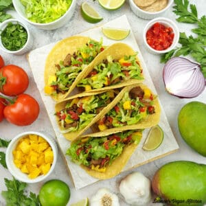 Seitan Tacos with bowls of toppings, mangos, garlic, onion, limes, and tomatoes