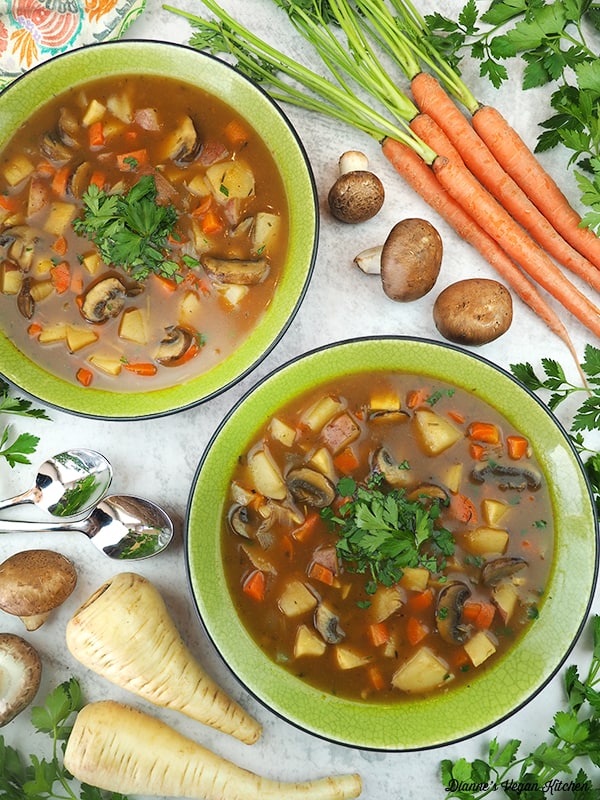 two bowls of vegan irish stew with spoons, parsnips, carrots, and mushrooms