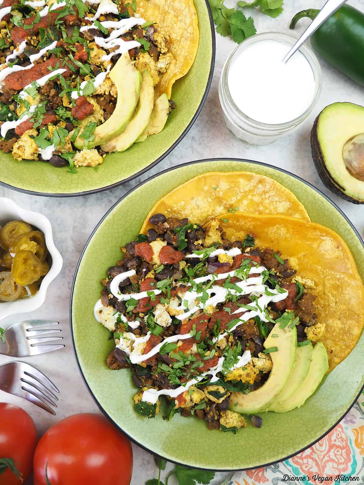 two plates of Vegan Huevos Rancheros with sour cream, tomatoes, jalapeños, and avocado