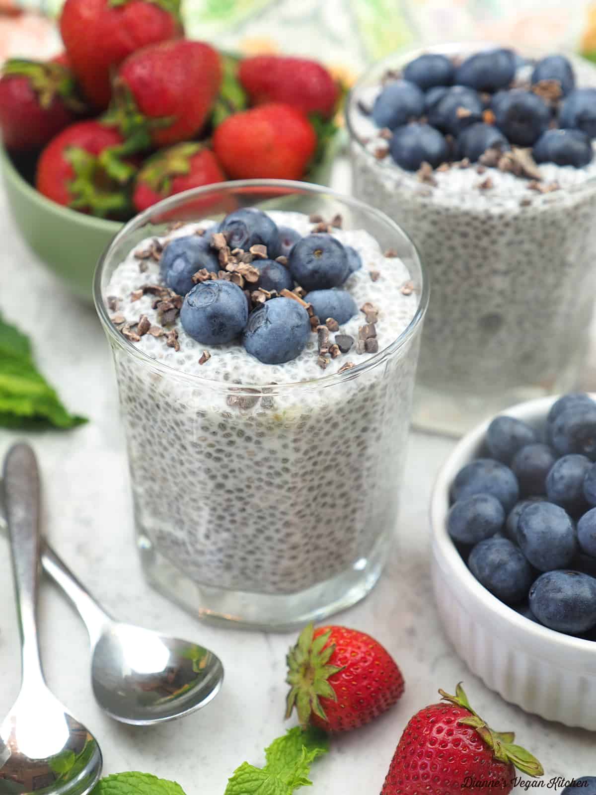 two chia puddings with a bowl of blueberries, spoons, and a bowl of strawberries