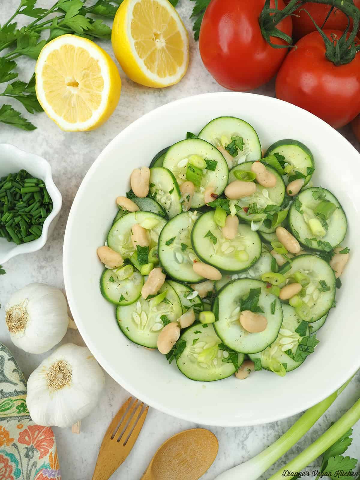 cucumber salad in a bowl surrounded by lemons, tomatoes, chives, garlic, a fork and spoon, and scallions