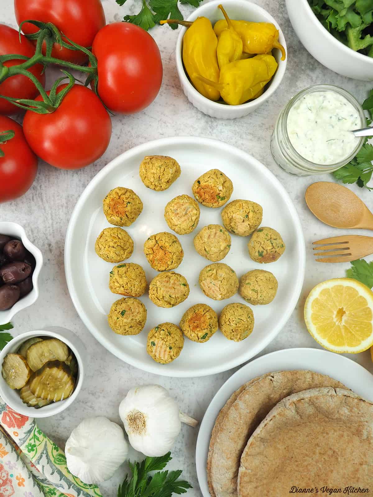 baked falafel balls on plate surrounded by peppers, tomatoes, sauce, lemons, garlic, olives, pickles, and pita bread