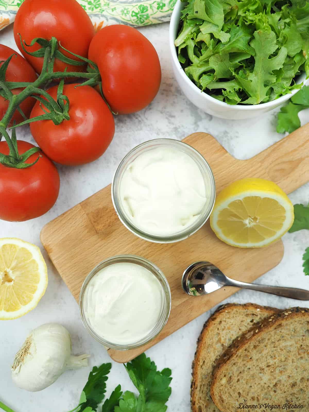 two jars of vegan mayonnaise overhead with a spoon, bread, garlic, lemons, salad, and tomatoes