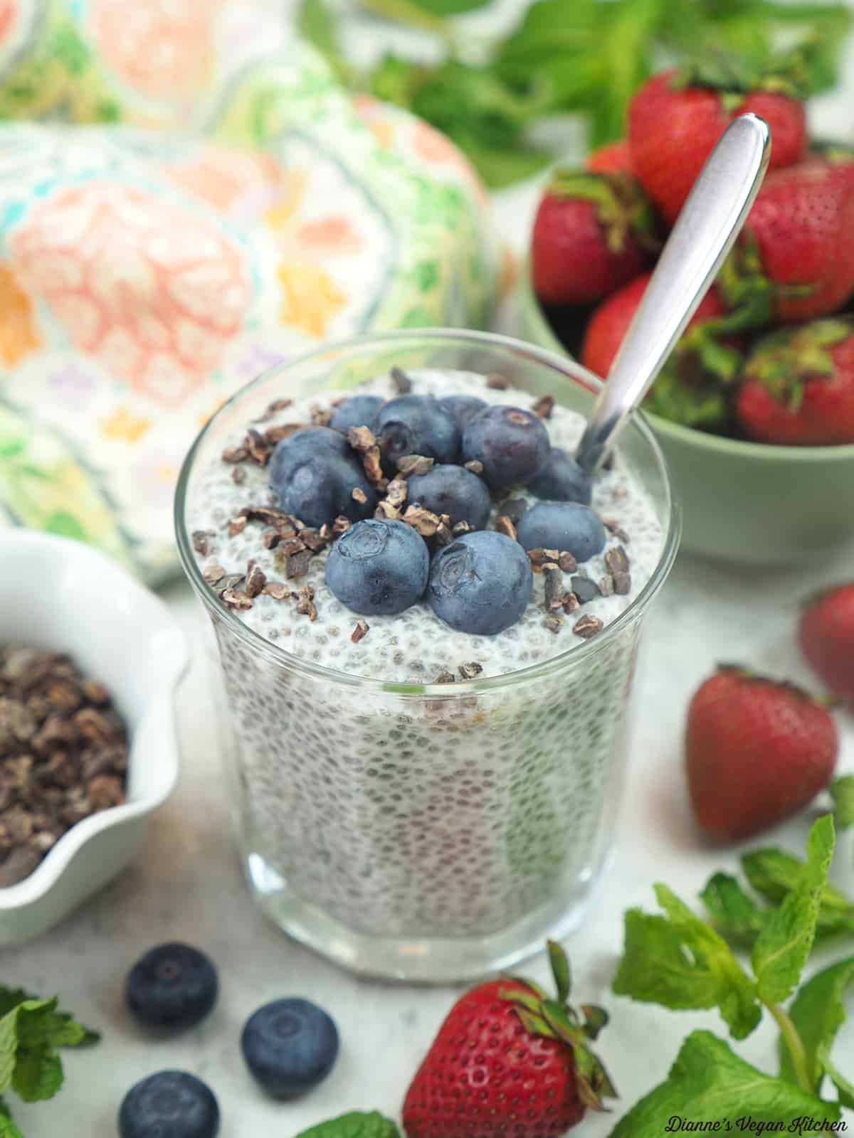 Blueberry Chia Pudding with a spoon, a bowl of strawberries, and a bowl of cacao nibs