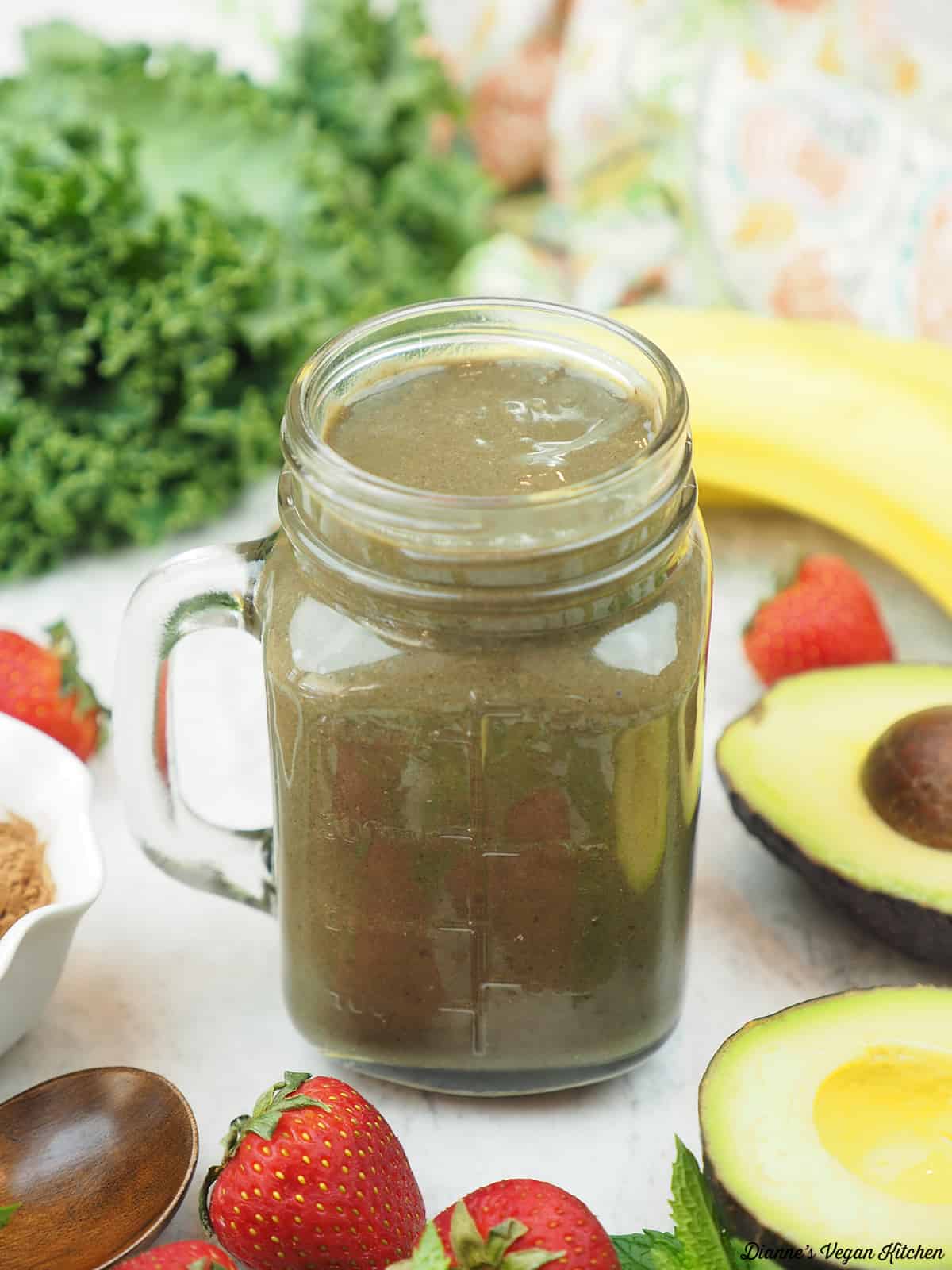 a chocolate shake surrounded by kale, an avocado, strawberries, bananas, and a bowl of cocoa powder