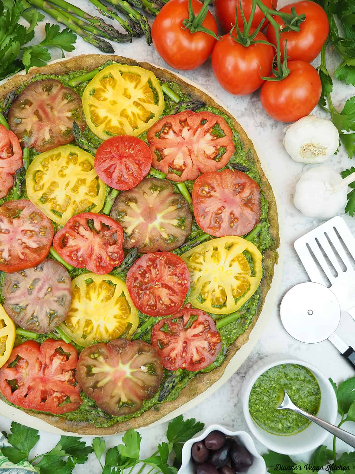 Grilled Pesto Tart surrounded by tomatoes, garlic, pesto, olives, and a pizza cutter