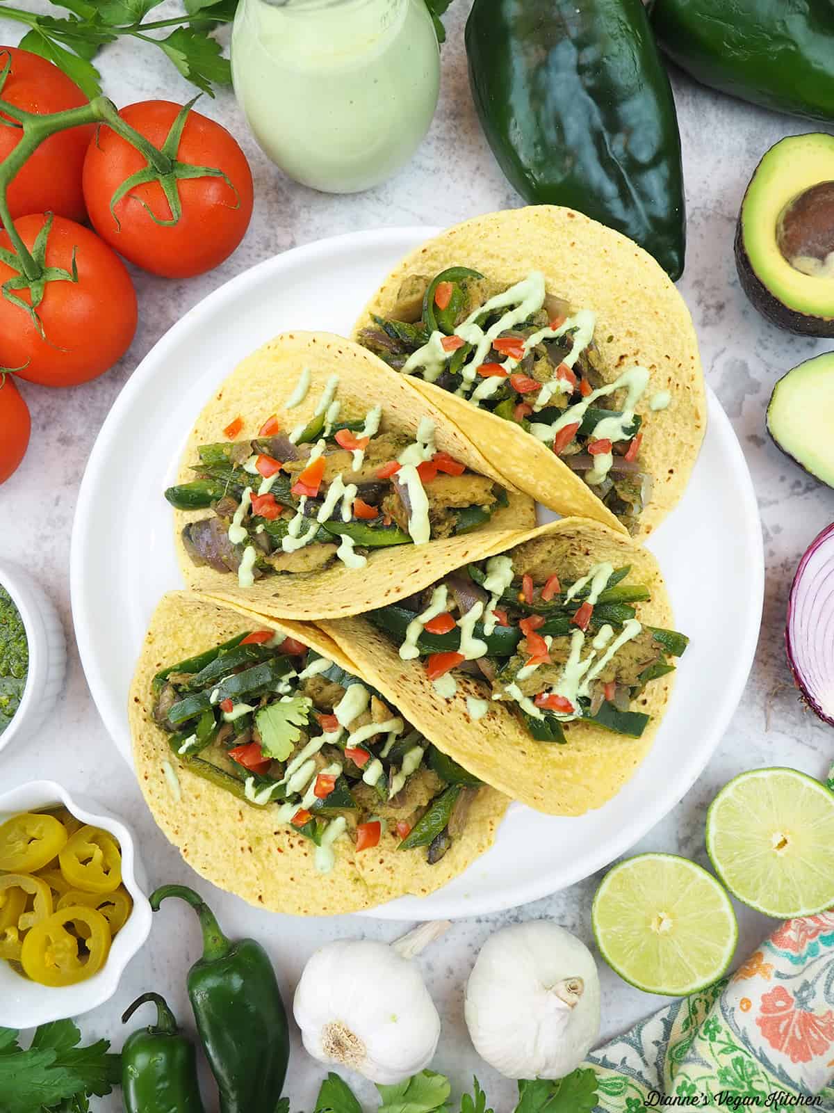 Seitan Chimichurri Tacos surrounded by tomatoes, jalapeño lime aioli, peppers, avocado, limes, garlic, onions, chimichurri sauce, and jalapeño slices